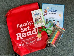 Picture of the summer reading challenge 2023 prizes: A red draw-string bag with the "Ready, Set, Read!" logo, a wooden medal, certificate, pack of coloured pencils and a folded leaflet.